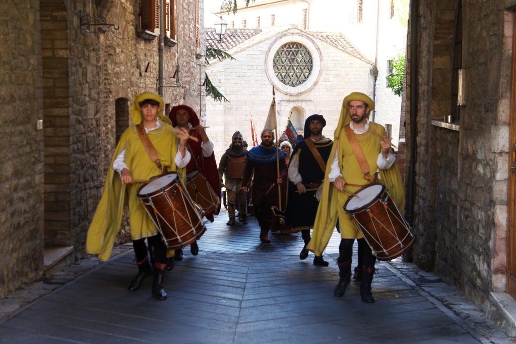 Gruppo di musicisti in costume storico che suonano tamburi mentre sfilano in una stretta via di un borgo medievale.