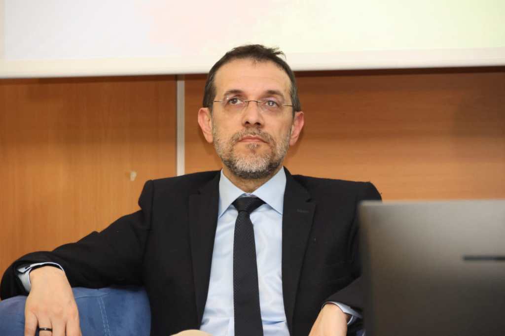 A man with glasses and a beard seated in a formal setting, wearing a suit and tie, looking contemplative while sitting next to a laptop.