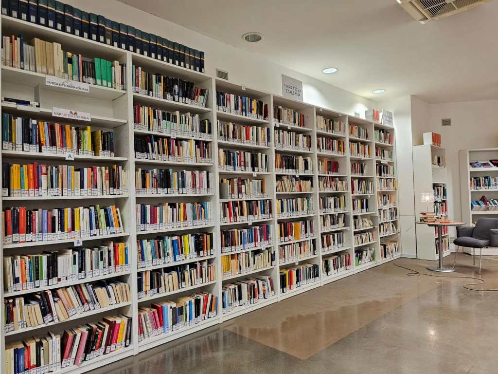 Interno di una biblioteca con scaffali bianchi pieni di libri disposti ordinatamente e un'area di lettura con poltrone e un tavolo.