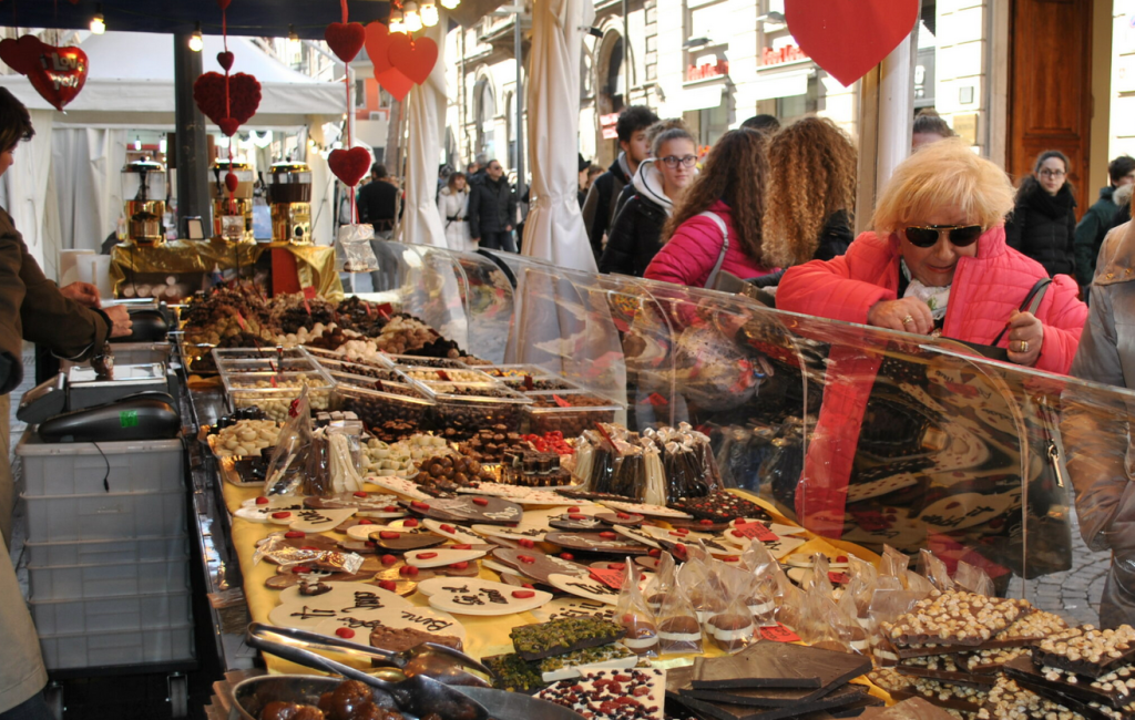 A bustling market scene featuring a wide array of chocolates and sweets displayed on a table, with a woman in a pink coat examining the treats. Colorful heart decorations hang above, and a crowd can be seen in the background.