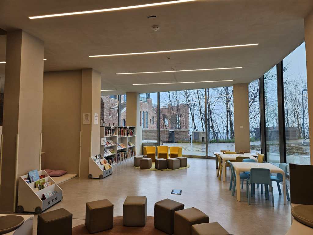 Interno moderno di una biblioteca con scaffali pieni di libri, tavoli e sedie colorate, grandi finestre che offrono luminosità e vista su alberi all'esterno.
