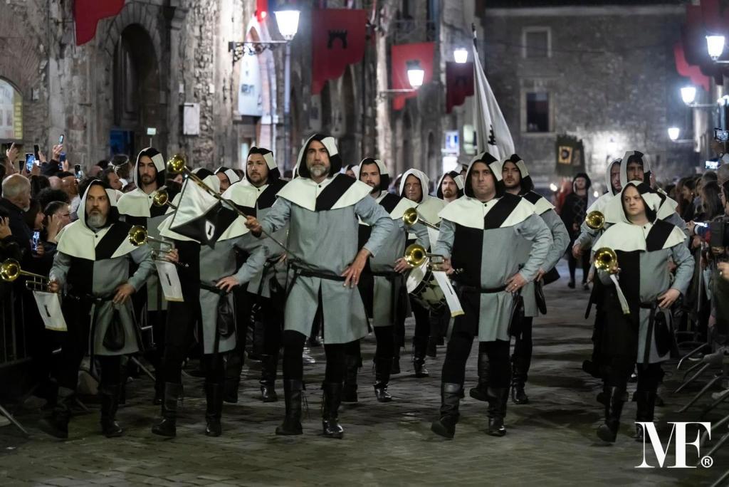 A group of men in medieval costumes, featuring gray and black outfits and hooded cloaks, parade down a cobblestone street during a festive event, with onlookers and banners in the background.