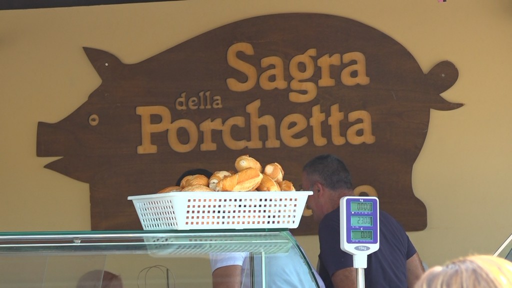 Insegna della Sagra della Porchetta di Costano con un maiale stilizzato, accanto a un venditore che serve panini in un negozio allestito per l'evento.