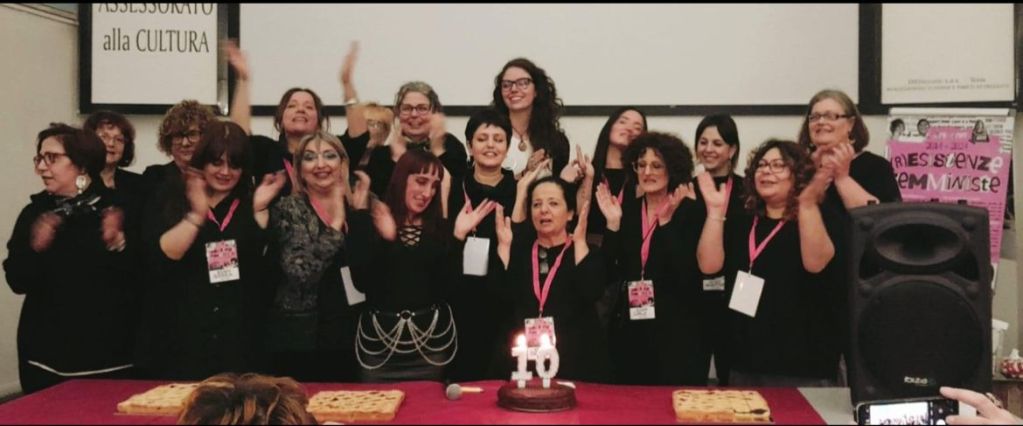 A group of women celebrating together, wearing black clothing, with some clapping and smiling. They are gathered around a table with a cake featuring the number 10, indicating an anniversary or milestone event.