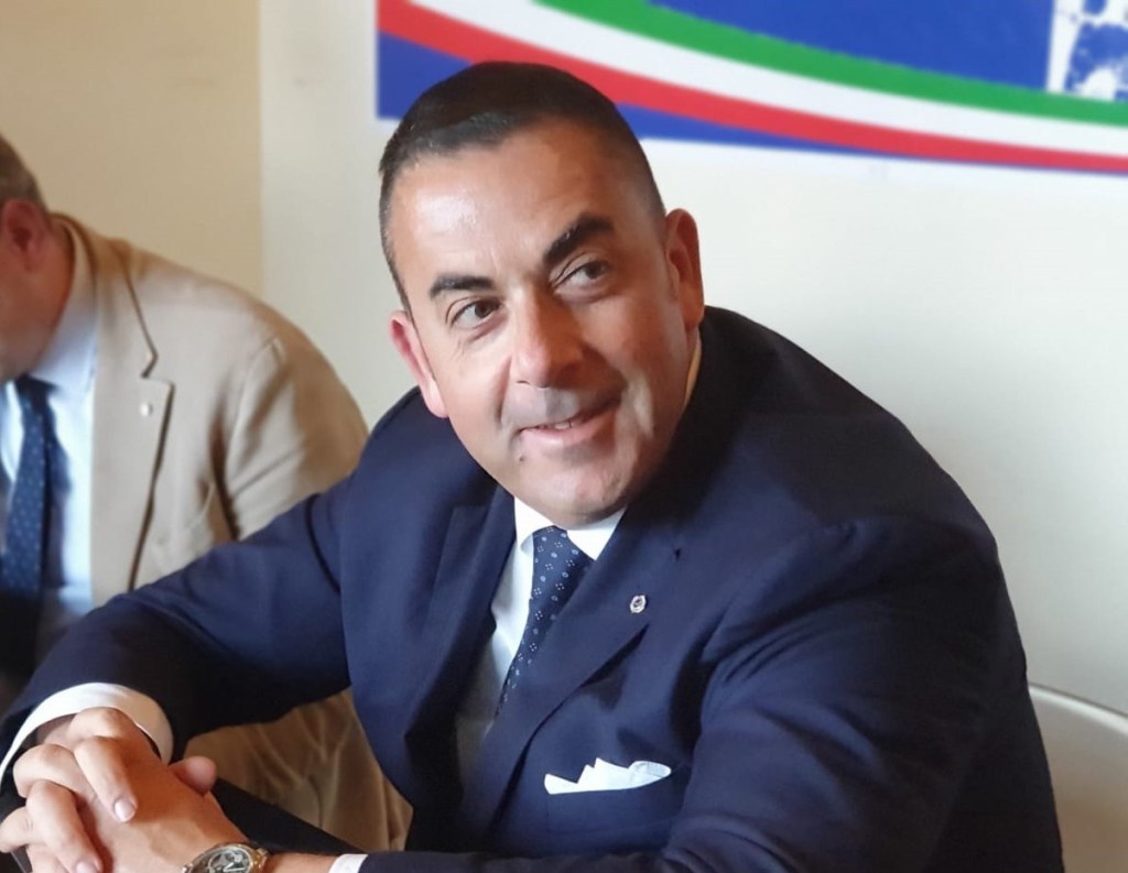 A man in a dark suit smiling while seated at a table with a light-colored background and a colorful flag behind him.
