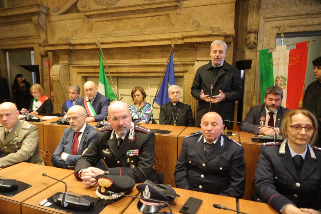A group of officials, including military personnel and local leaders, gathered at a round table in a formal setting, with national and regional flags displayed in the background.