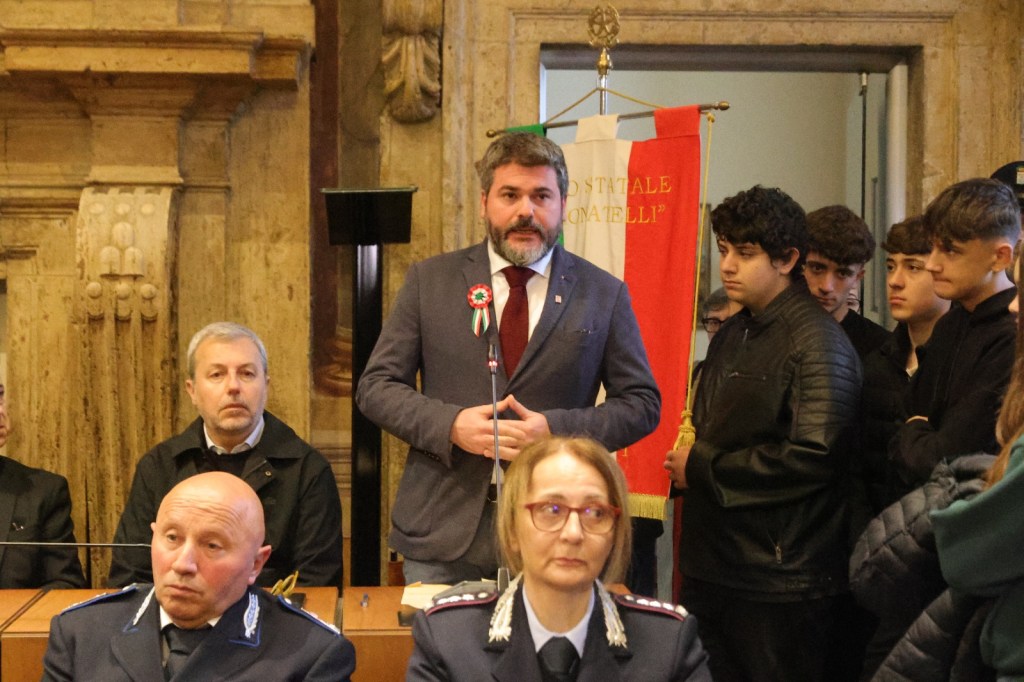 A group of people attending an event where a man is speaking. The man is wearing a suit and a rosette, with a flag in the background. Several individuals, including a woman in a police uniform, are seated and listening.