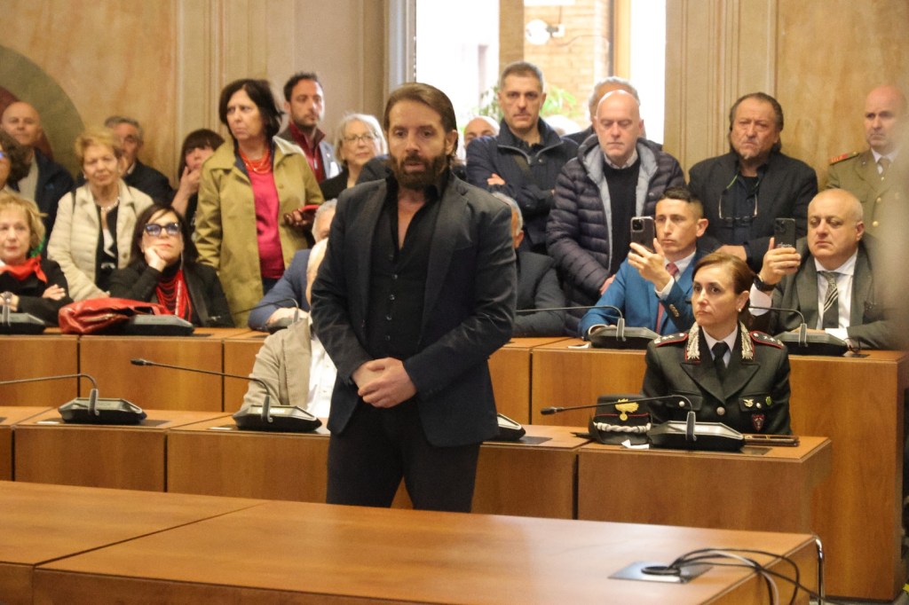 A man in a suit stands in front of a seated audience during a formal event, with various attendees, some holding smartphones, observing him. A woman in military attire is seated prominently at the front.