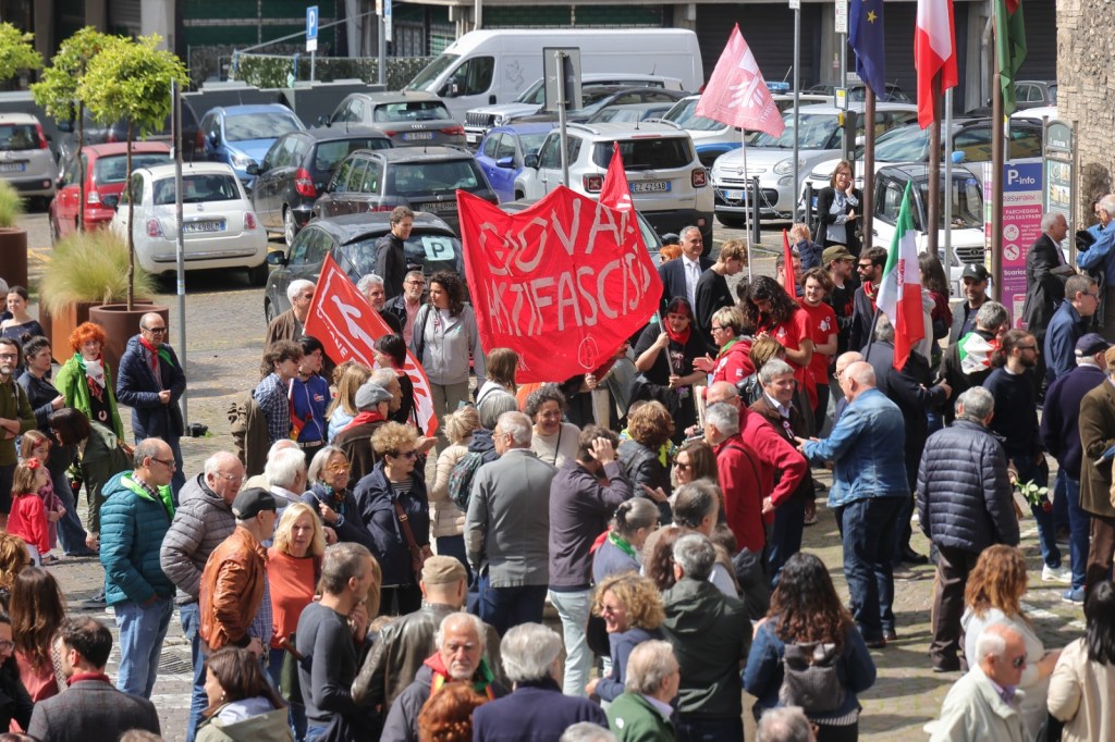 A large crowd gathers in an outdoor setting, holding flags and banners during a public demonstration, with various individuals wearing casual clothing.
