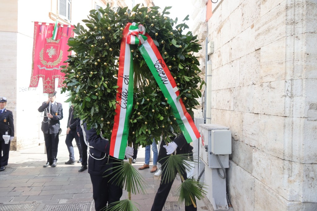 A ceremonial wreath made of greenery and decorated with ribbons in Italian colors, carried by a person in a formal uniform during a public event.