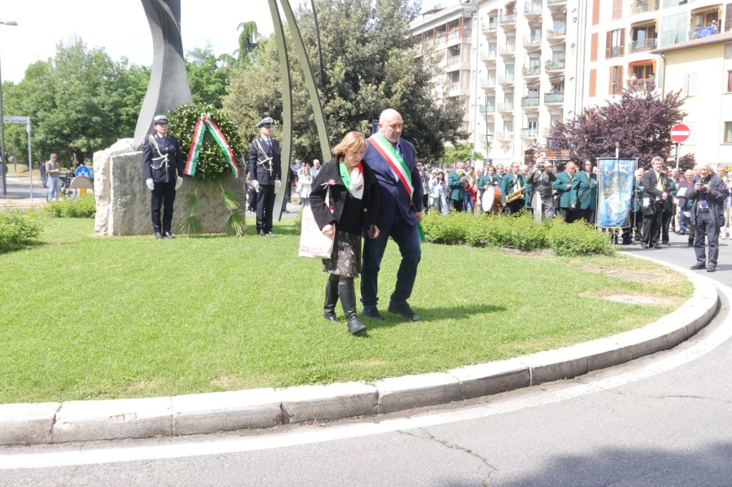 Two individuals walk together during an outdoor event, with one wearing a sash, while a crowd gathers in the background near a monument.