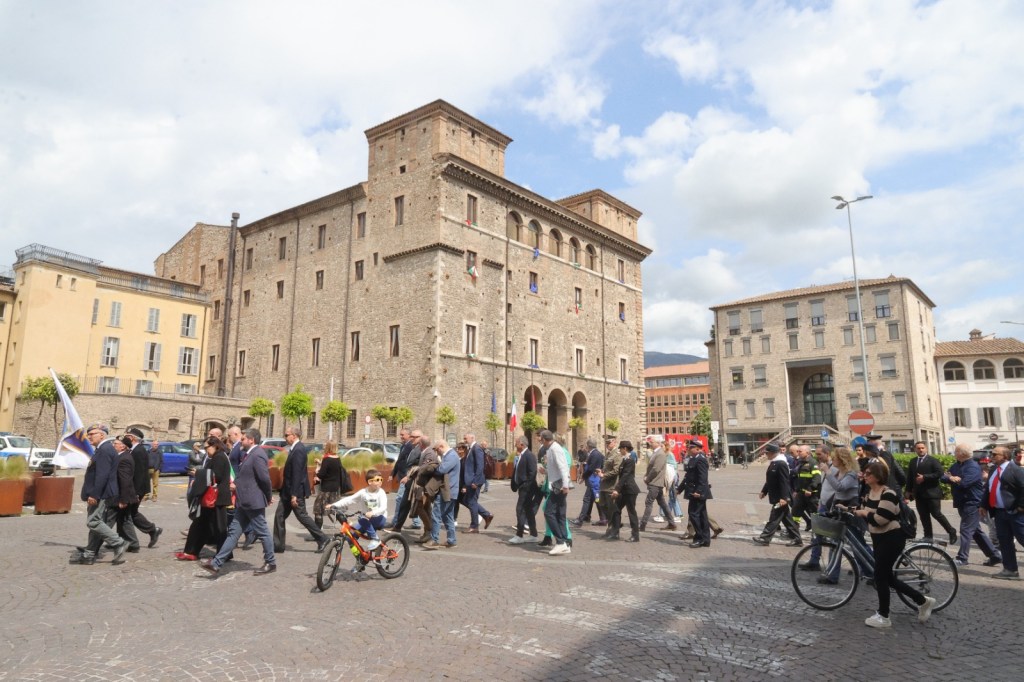 A crowd of people walking in a plaza, with an old stone building in the background and some modern buildings on the sides. A young boy on a bike is in the foreground, and the sky is partly cloudy.