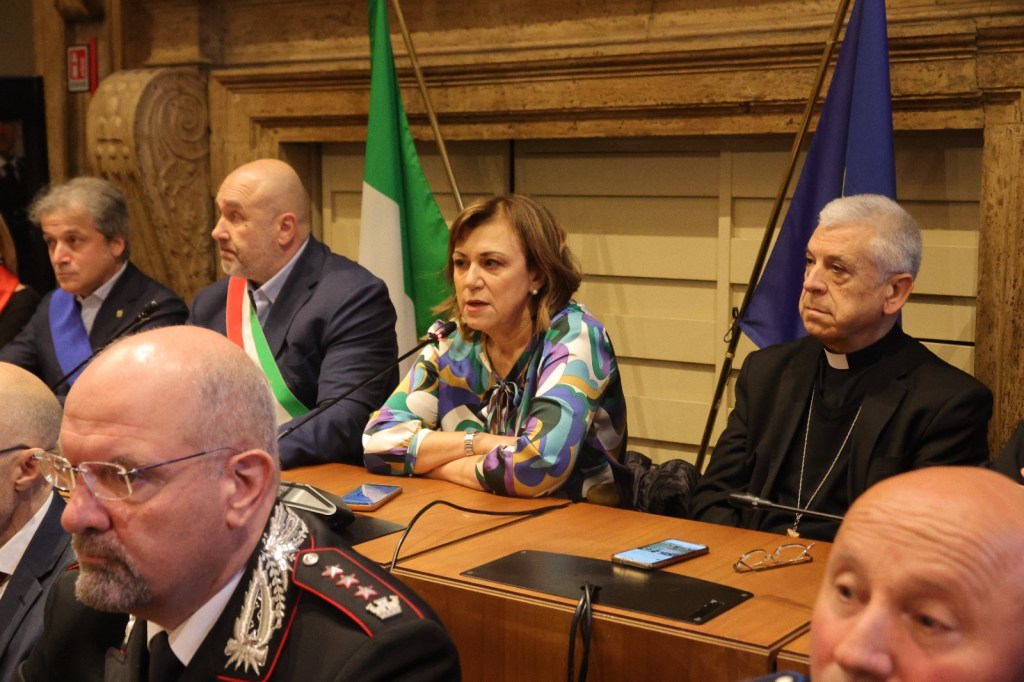 A group of officials seated at a table during a meeting, with Italian and European flags in the background. The officials include men and women in formal attire, with some wearing ceremonial sashes.