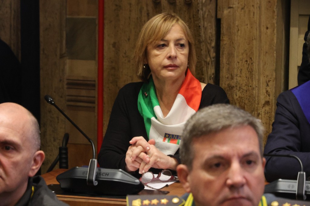 A woman wearing a white, green, and red scarf sits at a conference table, looking thoughtful. In the foreground, two men in military uniforms are also present.