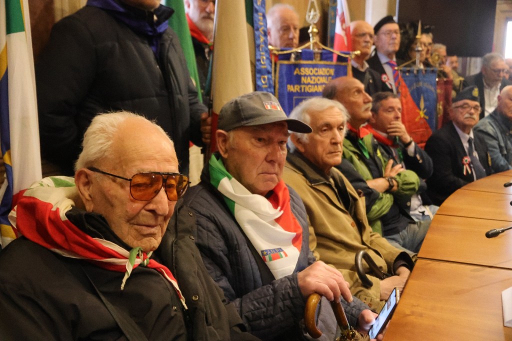 A group of elderly men attending a commemorative event, wearing scarves and hats, with banners in the background representing various organizations.