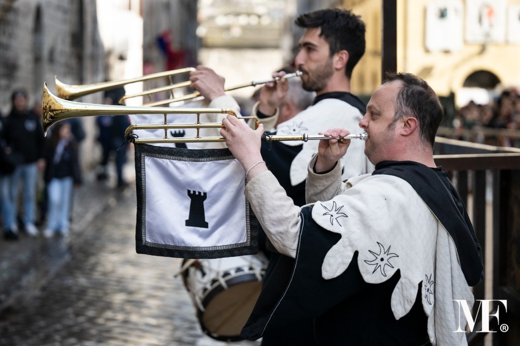 Musicisti con trombe vestiti in abiti storici durante una celebrazione a Narni, con bandiere e un pubblico di sfondo.