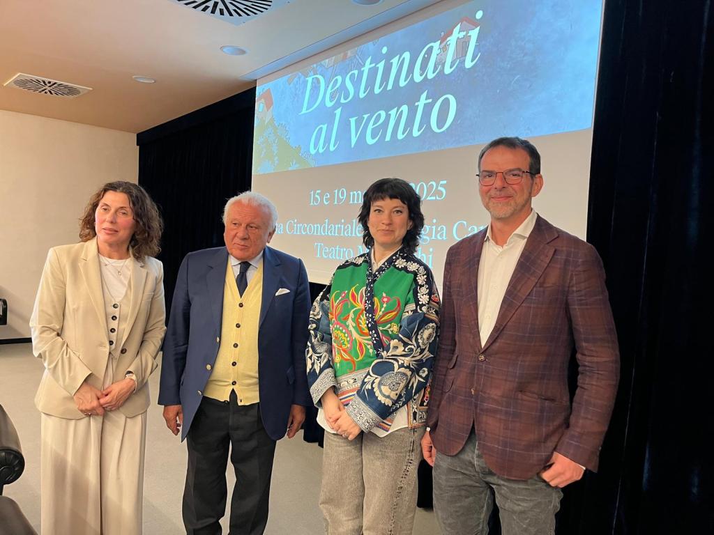 Un gruppo di quattro persone posa per una foto durante una conferenza stampa, con un grande schermo sullo sfondo che mostra il titolo dello spettacolo "Destinati al vento" e le date dell'evento.