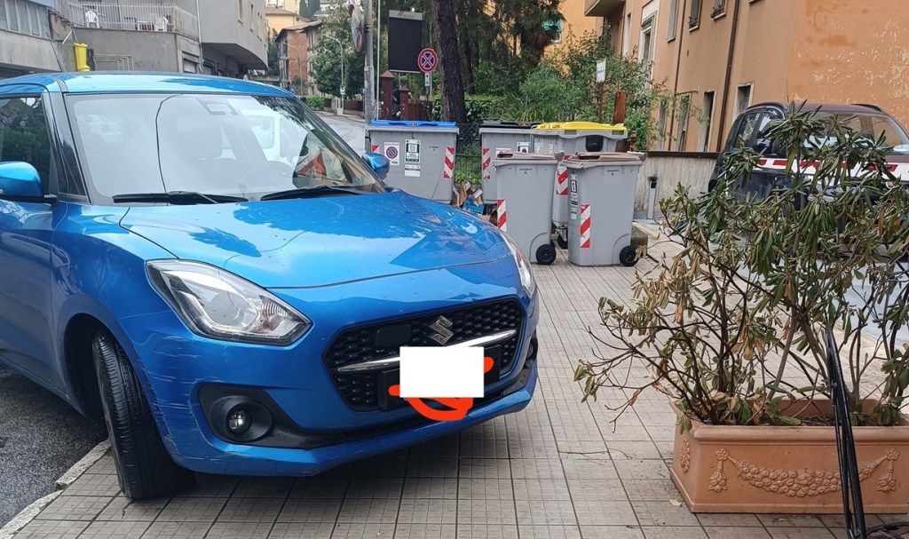 Un'auto blu parcheggiata sul marciapiede accanto a bidoni della spazzatura e una pianta in un quartiere di Perugia.