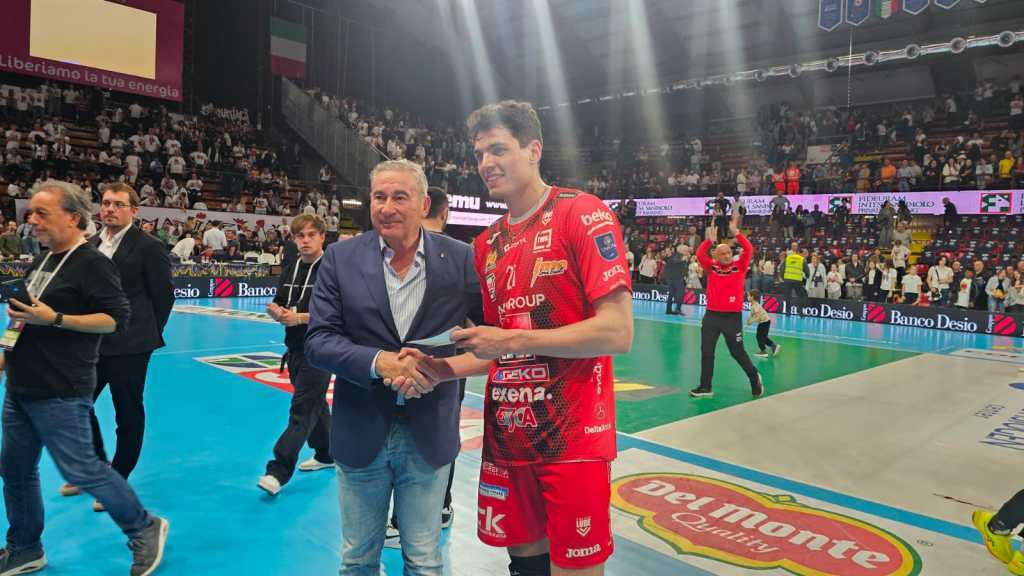 Giocatore di pallavolo premiato durante una cerimonia post-partita, mentre stringe la mano a un uomo in giacca blu, con il pubblico sullo sfondo.