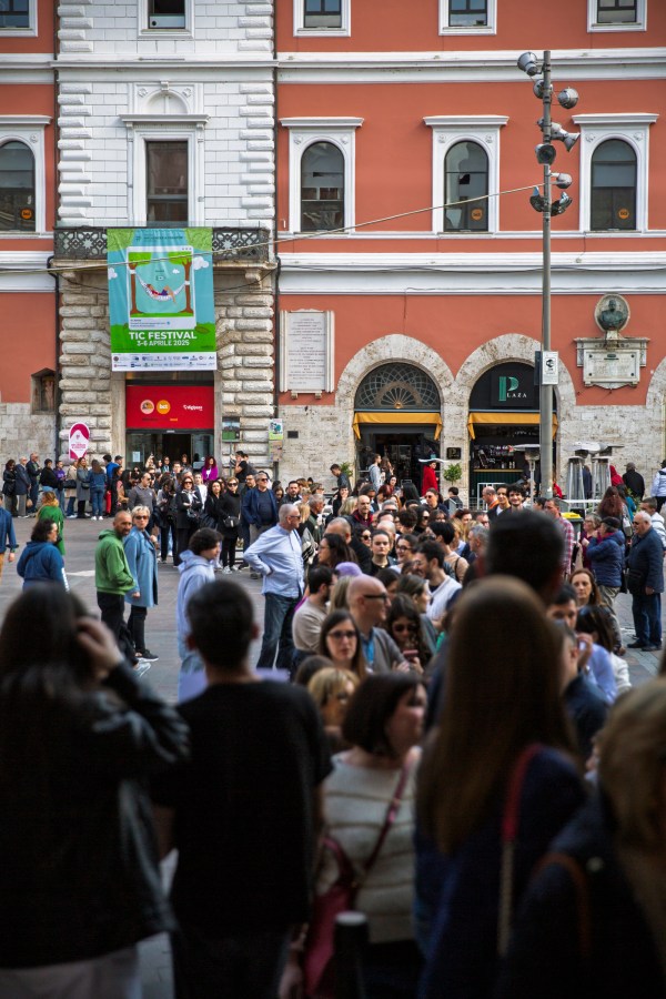 A crowded street scene with a large group of people gathered outside a building featuring a festival banner. Various individuals are seen in the foreground, with shops in the background.