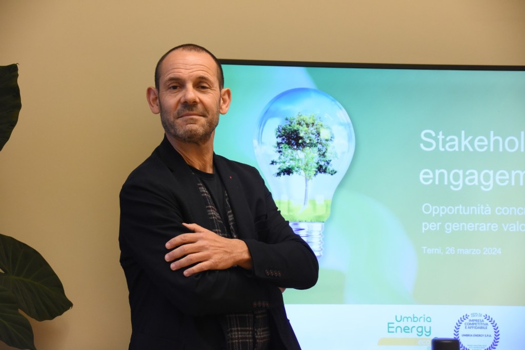 Un uomo con braccia incrociate sorride davanti a uno schermo che mostra un'immagine di una lampadina con un albero al suo interno, in un contesto aziendale legato a Umbria Energy.