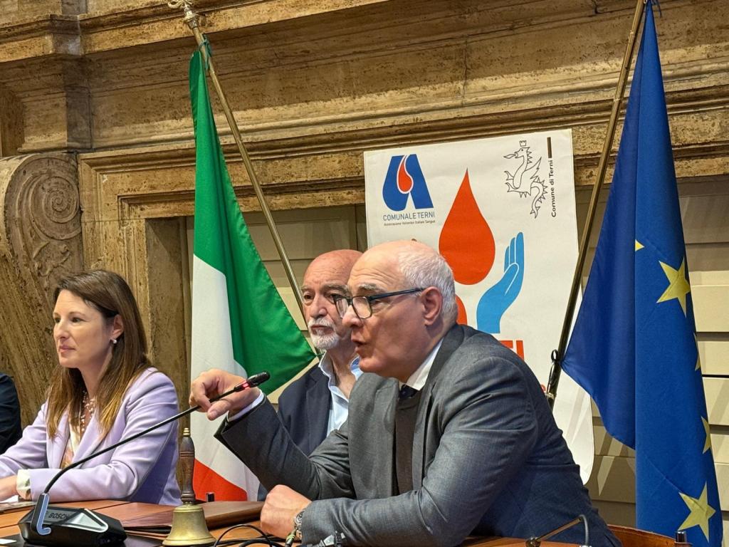 A meeting with three speakers at a podium: a woman in a light purple blazer, a man with a white beard, and another man in a gray suit, discussing in front of Italian and European Union flags and a backdrop featuring a logo.
