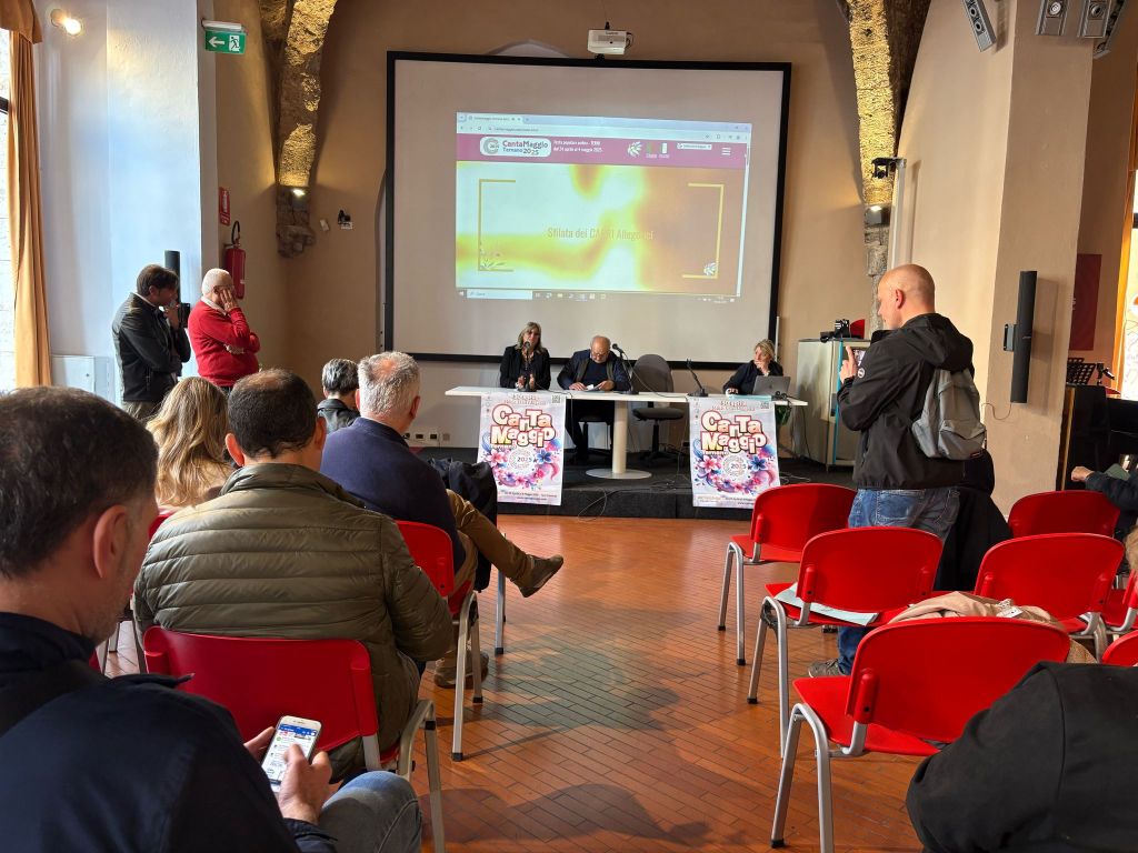 Una conferenza stampa riguardante la festa Cantamaggio a Terni, con diverse persone sedute in seggioline rosse e un relator presente al tavolo. Sullo sfondo, si vede uno schermo con informazioni sull'evento.