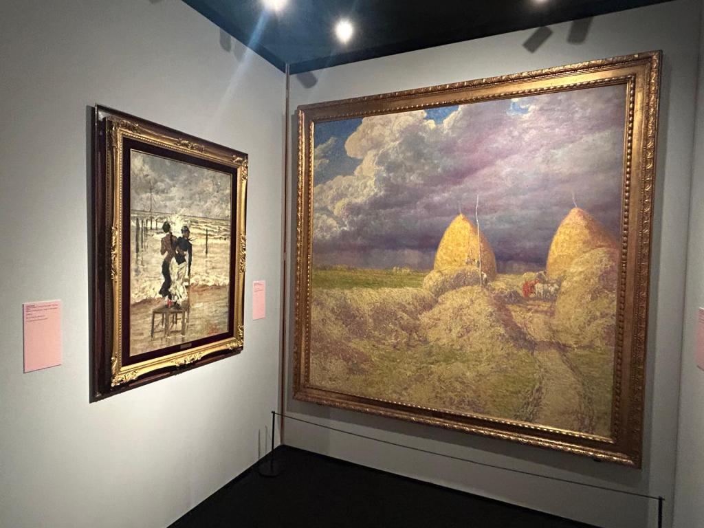 Due opere d'arte esposte in una galleria, una a sinistra con un dipinto che rappresenta una scena di vita quotidiana, e l'altra a destra raffigurante un paesaggio con fienili sotto un cielo nuvoloso.