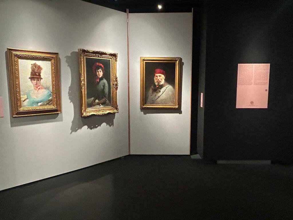 Tre dipinti esposti in una mostra d'arte, rappresentanti persone con abbigliamento storico, all'interno di una galleria con pareti chiare e scure.