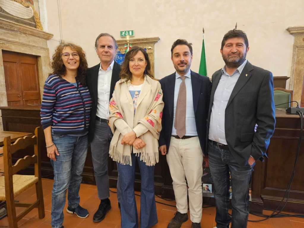 Gruppo di persone in piedi, sorridenti, durante una conferenza stampa nella Sala del Consiglio Comunale di Narni, con decorazioni murali e bandiere visibili sullo sfondo.