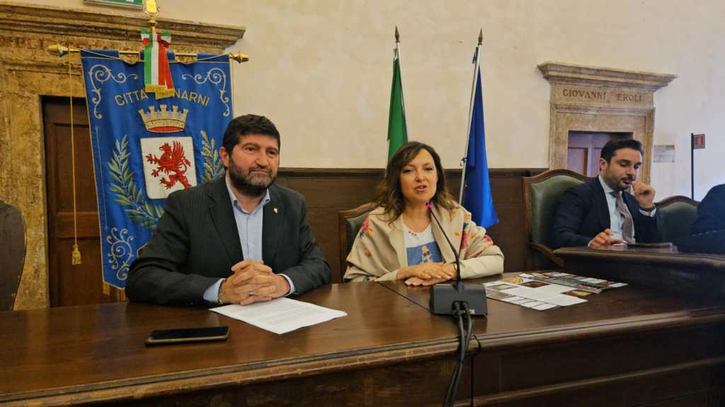 Due relatori seduti al tavolo durante una conferenza stampa, con una bandiera e il simbolo della città di Narni sullo sfondo.
