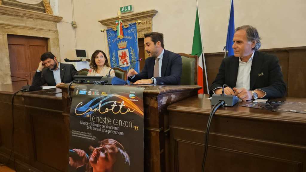 Conferenza stampa per il concerto di Carlotta al Teatro Manini di Narni, con vari relatori al tavolo, e un poster dell'evento in primo piano.
