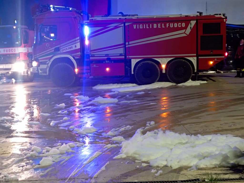 Autopompa dei vigili del fuoco con luci lampeggianti, di fronte a una superficie bagnata e schiumosa dopo un intervento d'emergenza.