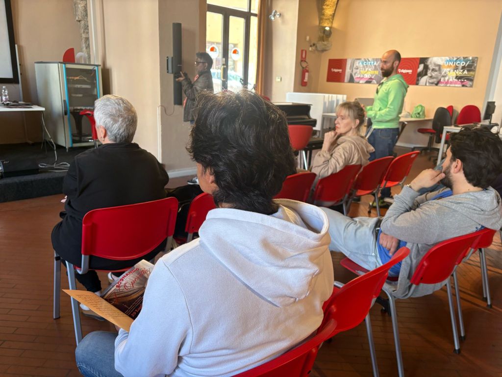 A group of people seated in a room, listening to a speaker. Some attendees are taking notes while others are facing the speaker, who is presenting in front of a screen.