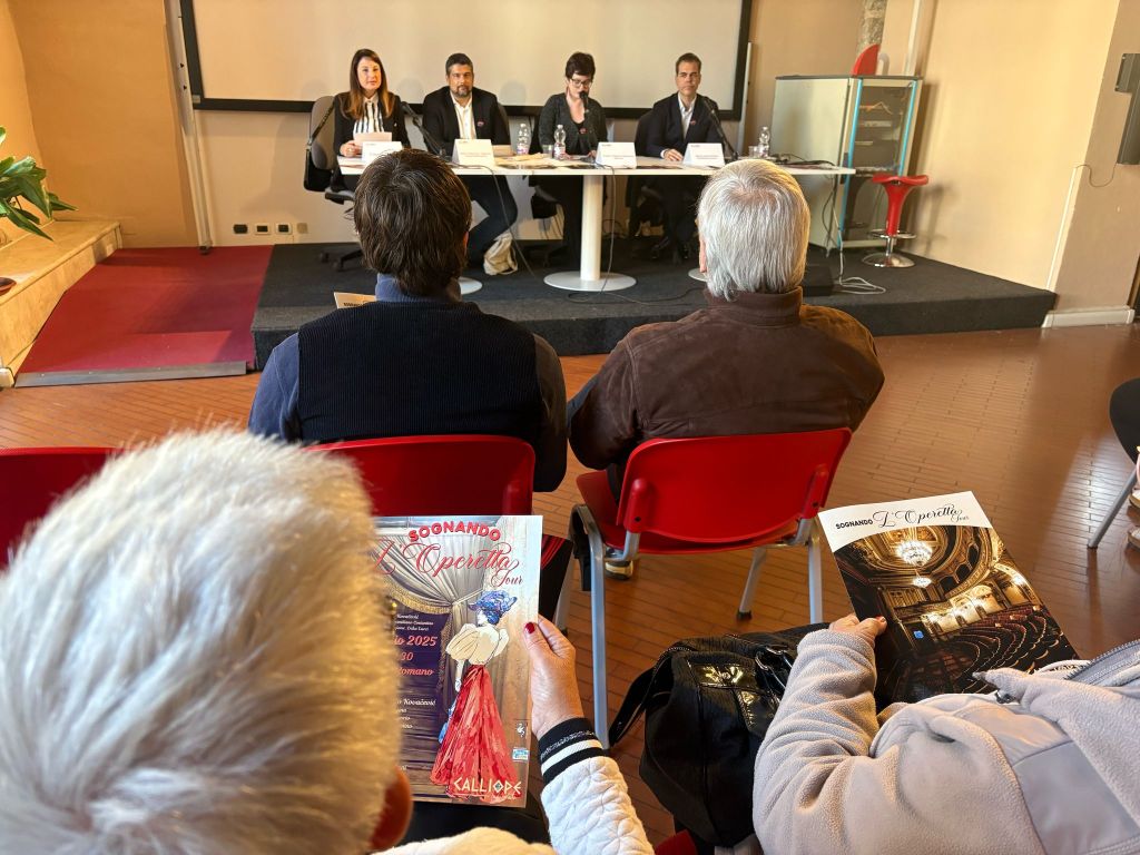 A panel discussion with four speakers sitting at a table, while an audience listens from chairs. Two attendees are holding brochures featuring vibrant colors and artwork.