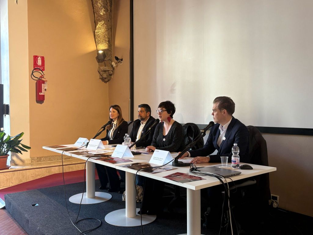 A panel of four speakers presenting at a conference, seated at a table with microphones and water bottles, in a well-lit room.