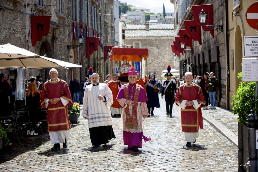 Processione religiosa con un vescovo e altre figure ecclesiastiche che camminano in una strada acciottolata durante le celebrazioni in onore di San Giovenale a Narni, con decorazioni e bandiere visibili.