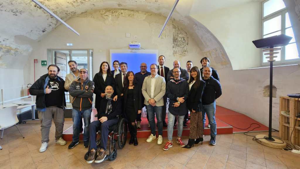 Un gruppo di persone sorridenti in una sala con pareti storiche, riunite per presentare la Corsa all'Anello di Narni. Sono presenti il sindaco e membri dell'amministrazione, insieme ad altri partecipanti.