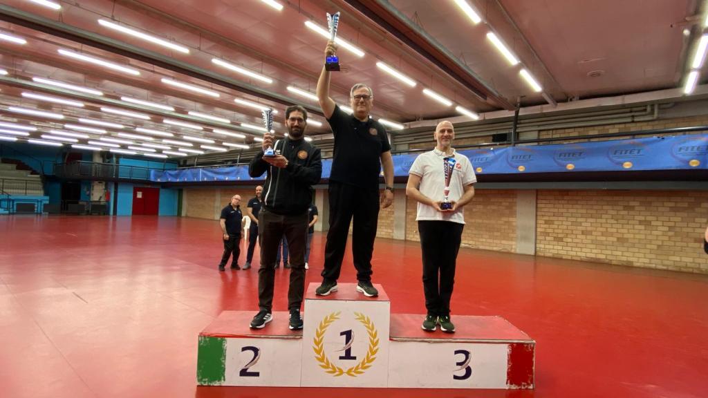 Tre atleti festeggiano sul podio del torneo di Wushu-Kung Fu, con il secondo e terzo classificato a sinistra e a destra, e il primo classificato al centro che tiene un trofeo.