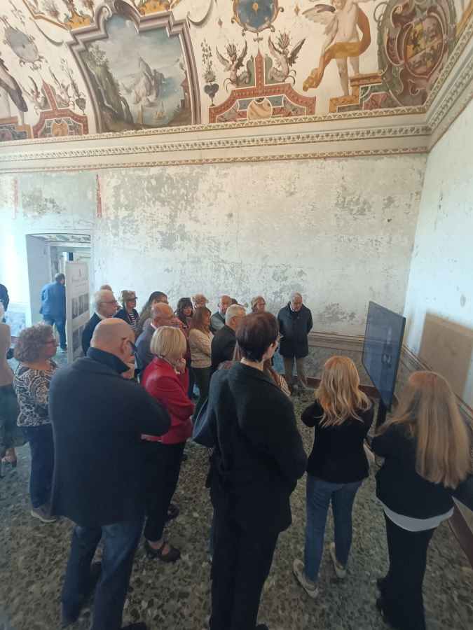 Un gruppo di persone radunate in una sala con affreschi sul soffitto, mentre osservano un display interattivo durante l'inaugurazione di una mostra dedicata a Alessandro Geraldini.