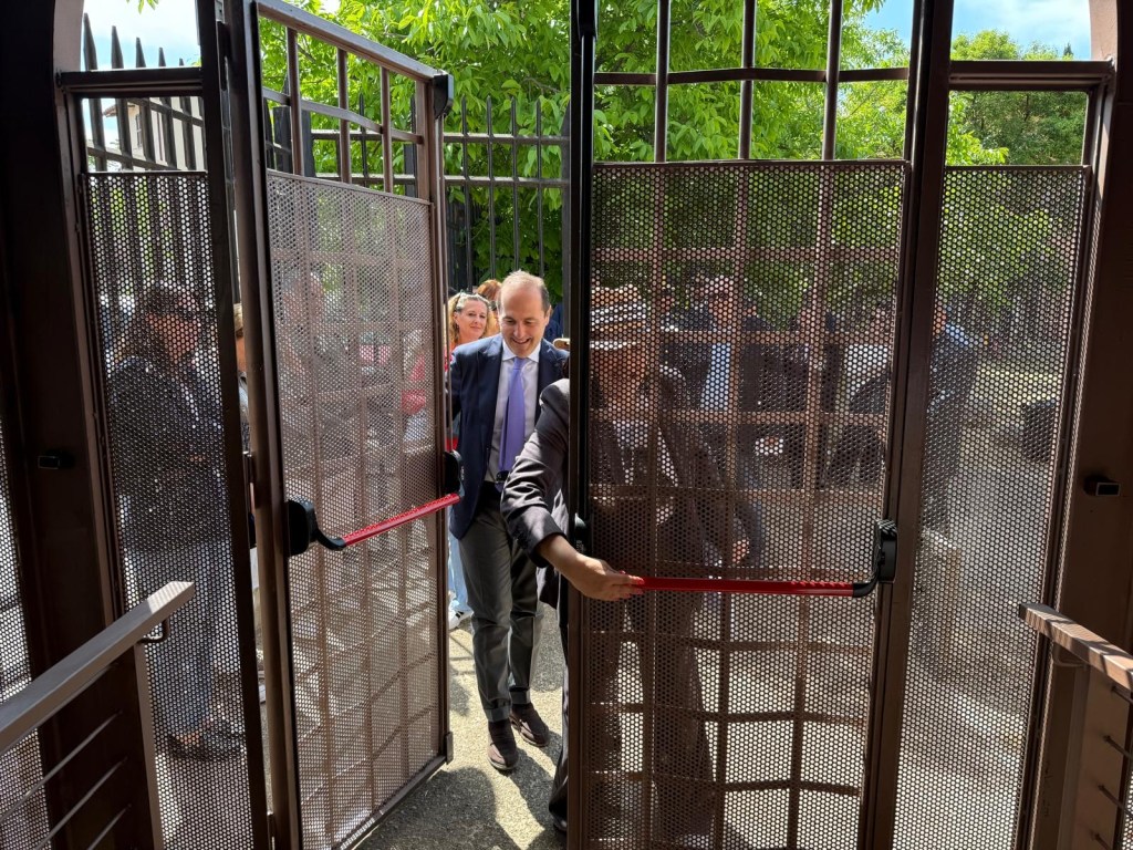 A group of people exiting through an open gate with metal mesh doors in a wooded area, with a man in a suit leading the way.