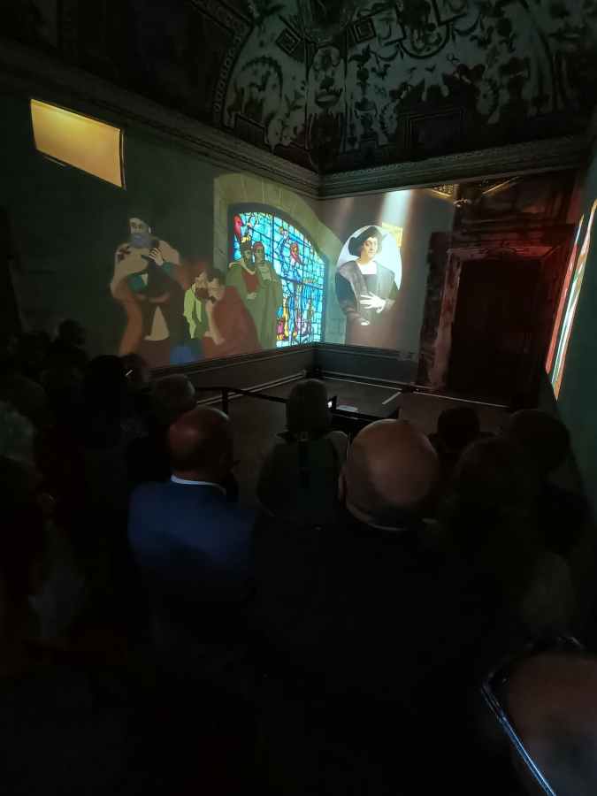 Un momento dell'inaugurazione della mostra multimediale su Alessandro Geraldini, con proiezioni artistiche su pareti e finestre ed un pubblico in ascolto.