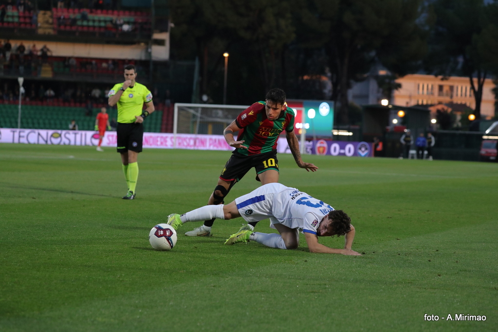 Giocatore in maglia rossa e verde tenta di recuperare la palla da un avversario a terra con maglia bianca, durante una partita di calcio.