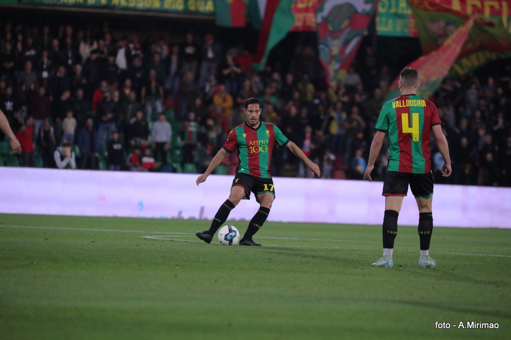 Giocatore in azione sul campo con una palla, mentre un compagno di squadra lo osserva, il pubblico sullo sfondo.