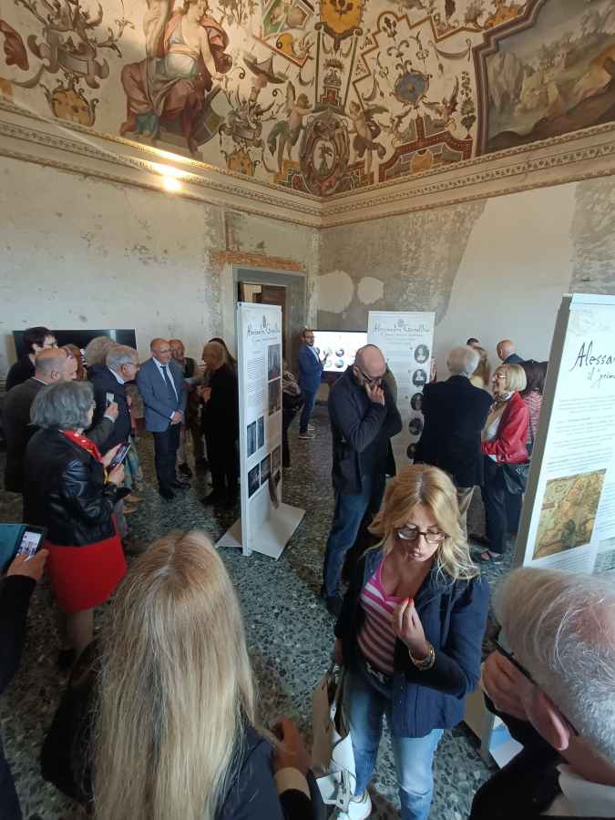 Una sala espositiva con un gruppo di persone che osservano pannelli informativi sulla vita di Alessandro Geraldini, circondati da affreschi storici.