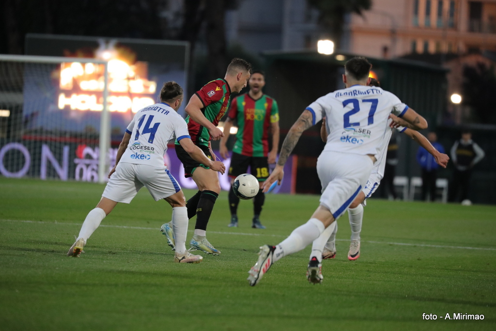 Un giocatore della Ternana con la maglia rossa e verde in azione mentre un avversario in maglia bianca tenta di fermarlo durante una partita di calcio.