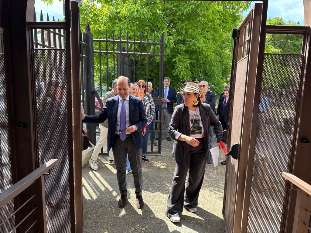 Two people entering through a large, open gate with a crowd visible in the background, surrounded by greenery.