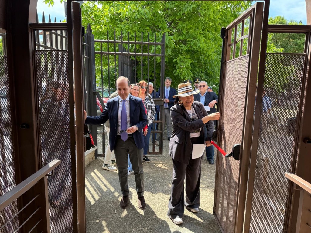 A group of people, including a man in a suit and a woman in a straw hat, are opening a large gate together. The scene is outdoors, set against a backdrop of greenery and other attendees.