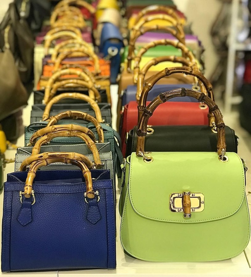 Una selezione di borse colorate con manici in bambù, esposte in riga su un tavolo, evidenziando la varietà di stili e colori disponibili per lo shopping.