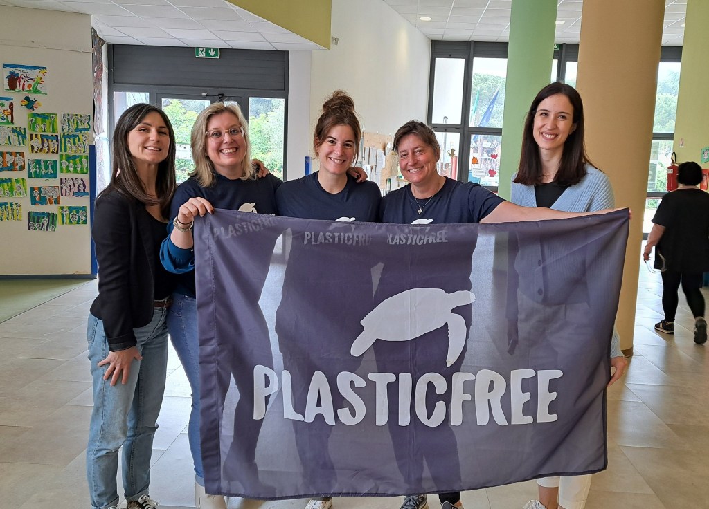 Five women hold a 'Plastic Free' banner inside a school, surrounded by colorful children's artwork on the walls.